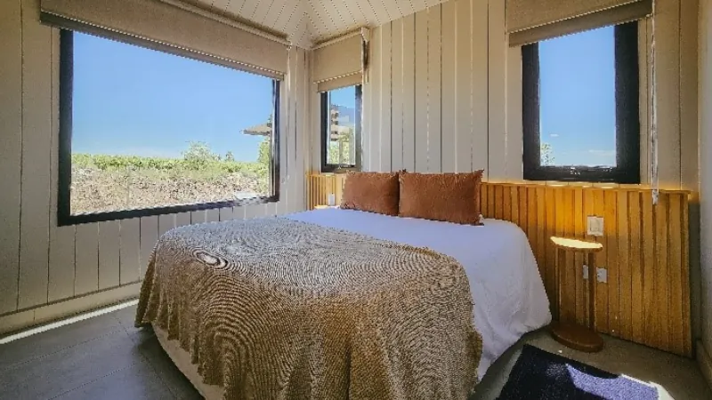 Bright bedroom with large windows, a double bed with brown pillows and a textured blanket, offering views of the surrounding landscape.