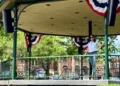 A decorated bandstand in Woodstock Town Square with patriotic bunting, surrounded by trees and historic brick buildings on a sunny day.