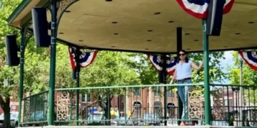 A decorated bandstand in Woodstock Town Square with patriotic bunting, surrounded by trees and historic brick buildings on a sunny day.
