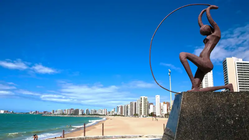 Bronze statue of a woman with raised arms holding a large circular arc, set on a pedestal by the beach in Fortaleza, Brazil, with high-rise buildings, sandy shoreline, and blue sky in the background.