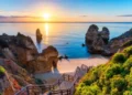 Sunrise over a rocky beach with cliffs and a wooden staircase leading down to the sand in Algarve, Portugal.