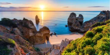 Sunrise over a rocky beach with cliffs and a wooden staircase leading down to the sand in Algarve, Portugal.