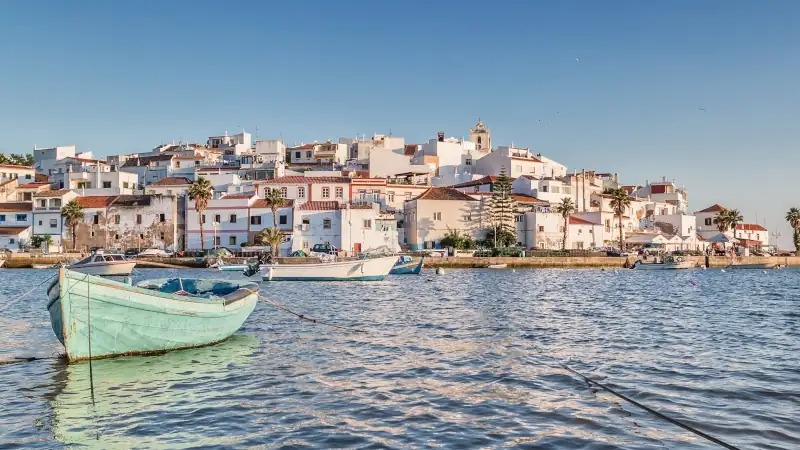 Boats floating on calm water in front of a coastal town with white buildings and red roofs under a clear sky.