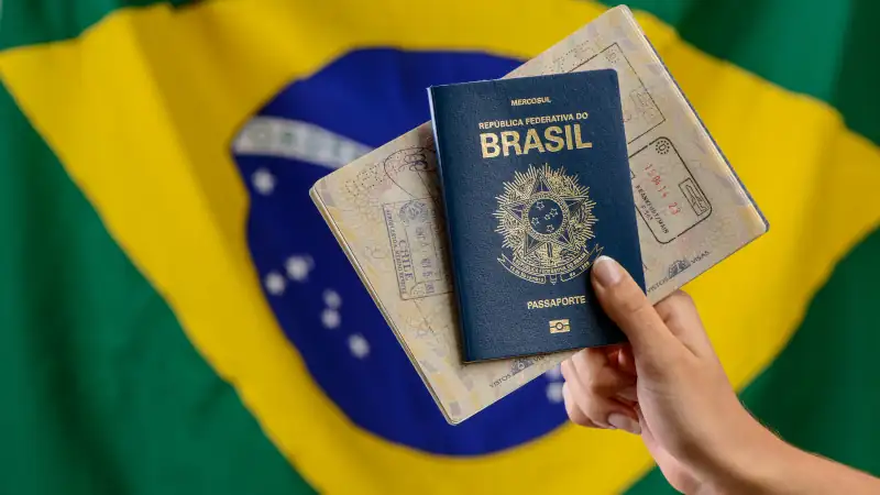 Hand holding Brazilian passport with Brazilian flag in the blurred background.