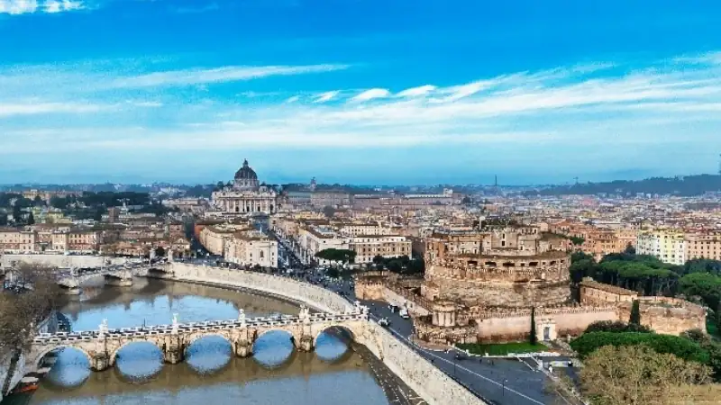 Aerial view of Rome with the Tiber River, Castel Sant'Angelo, and St. Peter's Basilica under a blue sky.