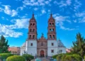 Front view of Durango Cathedral with twin bell towers under a bright blue sky, surrounded by green trees and a plaza.
