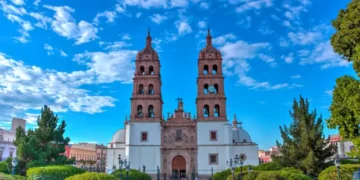 Front view of Durango Cathedral with twin bell towers under a bright blue sky, surrounded by green trees and a plaza.