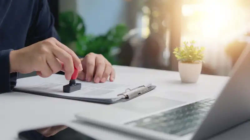 Individual in an office setting applies a stamp to a document contract legal agreement, signifying approval or verification certification permit visa next to an open laptop.