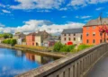 Colorful riverside buildings in Kilkenny, Ireland, viewed from a stone bridge under a blue sky.