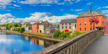 Colorful riverside buildings in Kilkenny, Ireland, viewed from a stone bridge under a blue sky.