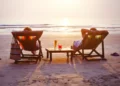 Two people relax in beach chairs facing the ocean at sunset, with drinks on a small table between them.