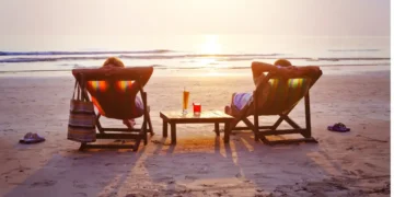 Two people relax in beach chairs facing the ocean at sunset, with drinks on a small table between them.
