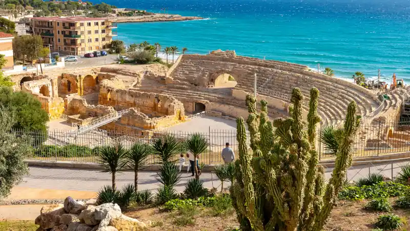 Roman amphitheater in Tarragona, Spain, overlooking the Mediterranean Sea with cactus and palm trees in the foreground.
