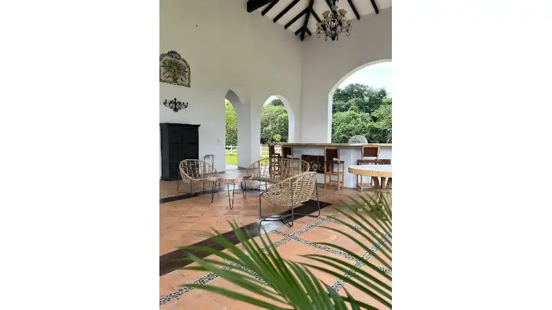 Open-air social area with wicker chairs, tiled floor, arched windows, and a bar overlooking lush greenery.