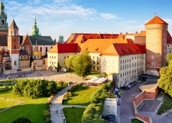 A panoramic view of Wawel Castle in Kraków, Poland, showcasing its red-roofed buildings, cathedral towers, and a landscaped courtyard bathed in daylight.