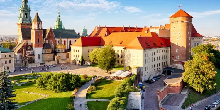 A panoramic view of Wawel Castle in Kraków, Poland, showcasing its red-roofed buildings, cathedral towers, and a landscaped courtyard bathed in daylight.