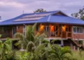 A raised wooden house with a blue roof and solar panels, surrounded by tropical vegetation.