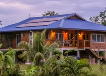 A raised wooden house with a blue roof and solar panels, surrounded by tropical vegetation.