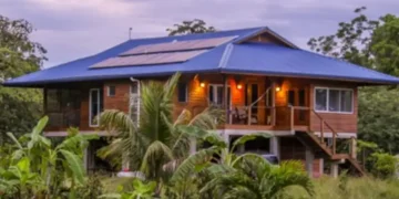 A raised wooden house with a blue roof and solar panels, surrounded by tropical vegetation.