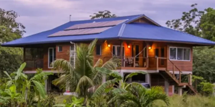 A raised wooden house with a blue roof and solar panels, surrounded by tropical vegetation.