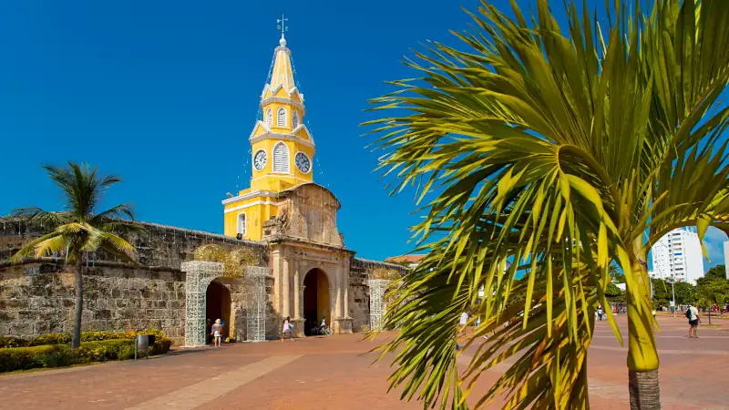 Historic entrance gate of Cartagena, Colombia, with a yellow clock tower, palm trees, and bright blue skies.