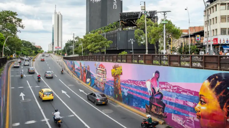 Colorful mural on a city wall in Cali, Colombia, depicting cultural and urban scenes along a busy road with cars and motorcycles.