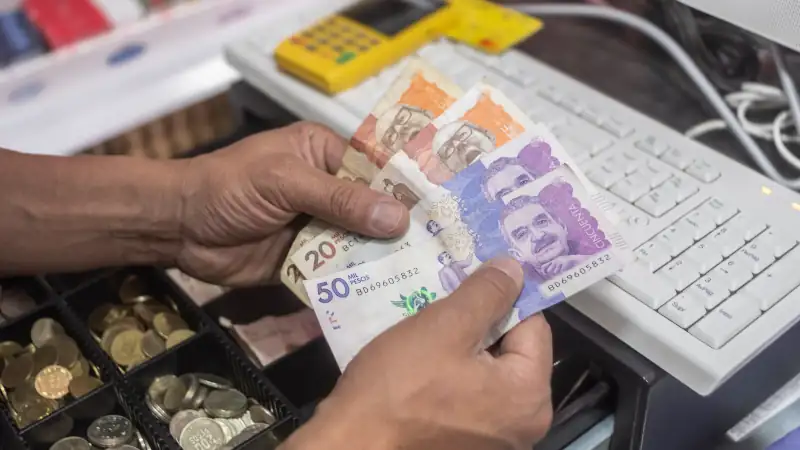 Person handling Colombian peso banknotes at a cash register with coins and a keyboard nearby.