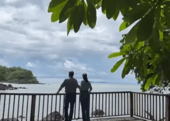 Lief and Kathleen standing together on a wooden viewing deck, gazing out over a calm ocean surrounded by lush greenery and a cloudy sky.