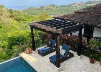 Tropical patio with lounge chairs, pool, lush greenery, and mountain view. One person relaxing.