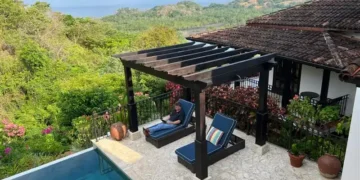 Tropical patio with lounge chairs, pool, lush greenery, and mountain view. One person relaxing.