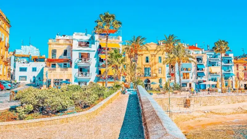 Colorful seafront buildings and palm trees along a sunny promenade in Sitges, Spain.