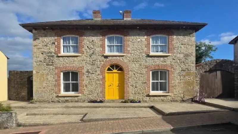 A two-story stone house with red brick trim around the windows and a bright yellow front door, under a blue sky with some clouds.