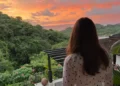 Kathleen Peddicord stands on a terrace, facing away from the camera, overlooking a lush green landscape and a vibrant orange-pink sunset sky.