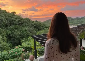Kathleen Peddicord stands on a terrace, facing away from the camera, overlooking a lush green landscape and a vibrant orange-pink sunset sky.