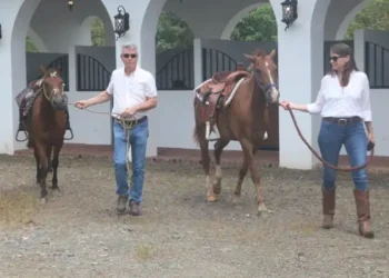 Lief and Kathleen are leading two saddled horses outside a white-arched stable. Both are dressed in white shirts and jeans, walking on a gravel path at an equestrian facility.