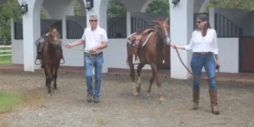 Lief and Kathleen are leading two saddled horses outside a white-arched stable. Both are dressed in white shirts and jeans, walking on a gravel path at an equestrian facility.