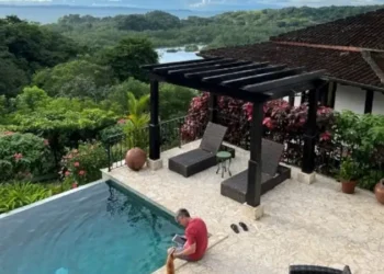 Lief Simon sitting by an infinity pool overlooking a lush tropical forest and distant water view, with lounge chairs under a pergola on a stone patio.