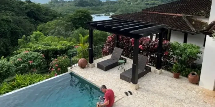 Lief Simon sitting by an infinity pool overlooking a lush tropical forest and distant water view, with lounge chairs under a pergola on a stone patio.