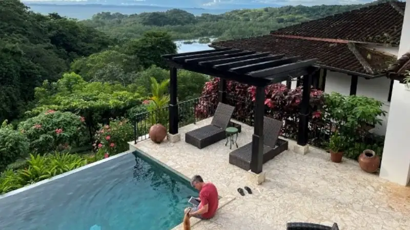 Lief Simon sitting by an infinity pool overlooking a lush tropical forest and distant water view, with lounge chairs under a pergola on a stone patio.