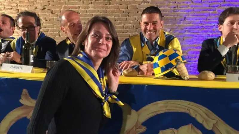 Kathleen stands smiling in front of a table of men in yellow and blue scarves during a festive indoor event, likely related to the Palio di Siena.