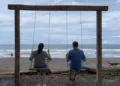 Lief and Kathleen swinging on a wooden swing set by the beach, facing the ocean with waves and palm trees in the background.