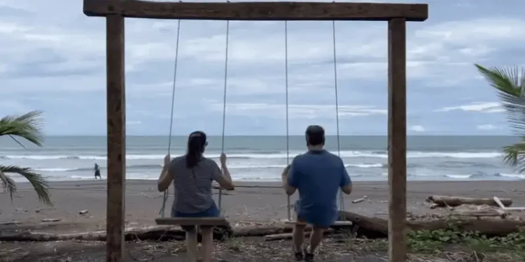 Lief and Kathleen swinging on a wooden swing set by the beach, facing the ocean with waves and palm trees in the background.