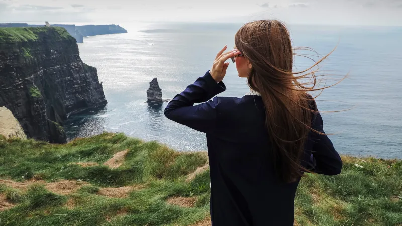 Young and stylish girl tourist posing with stunning Cliff of Moher in the background. County Clare, Ireland. Travel and tourism. Exploring famous Irish landmark.