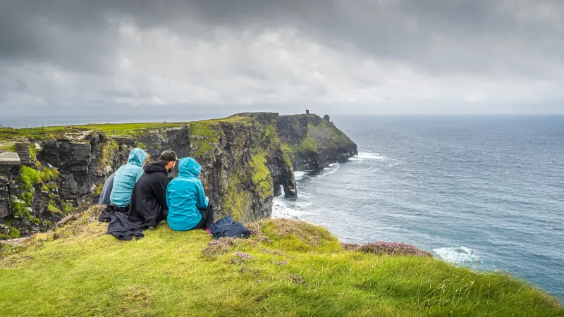 Group of young people sitting on the edge of cliff on iconic Cliffs of Moher, popular tourist attraction, Wild Atlantic Way, County Clare, Ireland