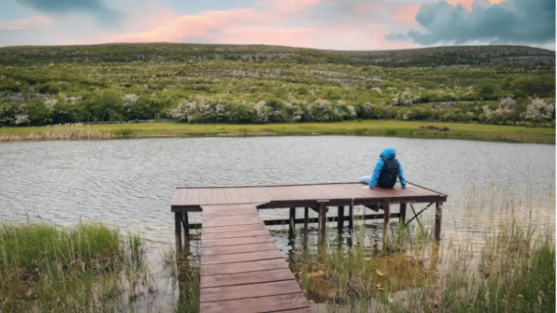 Hiker in blue jacket sitting on wooden pier by the lake with mountain in background at Burren national park, county Clare, Ireland, Adventure and lifestyle concept, wallpaper