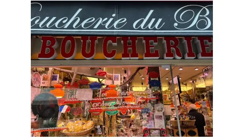 French butcher shop storefront with colorful displays, hanging lanterns, and a customer browsing inside.