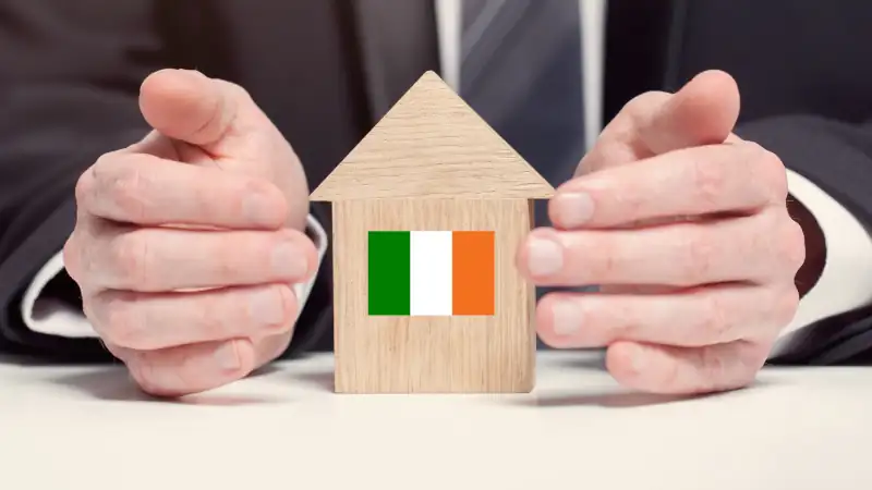 Businessman hand holding wooden home model with Irish flag