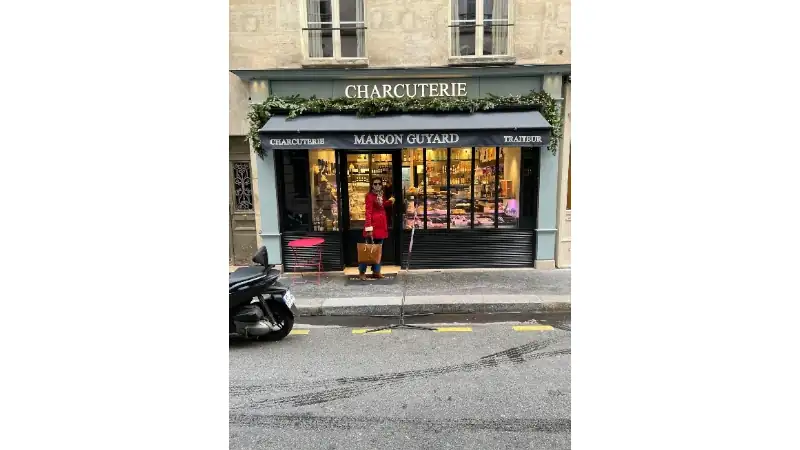 Kathleen standing outside a Parisian charcuterie shop with a bag, in front of the storefront ‘Maison Guyard’ on a city street.
