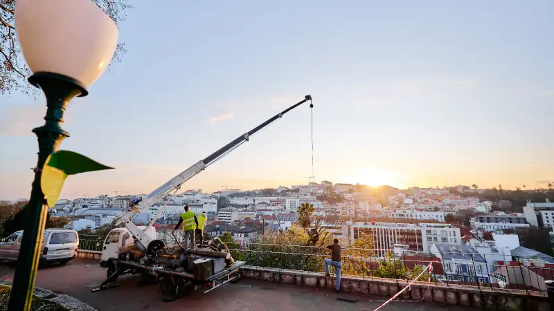 Workers operating a crane on a hilltop overlooking a cityscape at sunset.