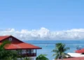 Panoramic view of Bocas del Toro Bay with red-roofed waterfront homes, palm trees, and sailboats anchored on calm tropical waters under a bright blue sky.